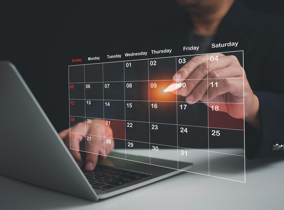 Hand marking dates on a digital calendar above a laptop